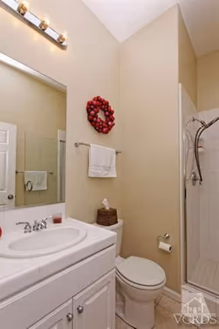 A clean bathroom with a white sink vanity, a large mirror above the sink, a toilet next to the vanity, a towel rack with a white towel, a decorative red wreath on the wall, and a glass-enclosed shower with a curved shower rod.