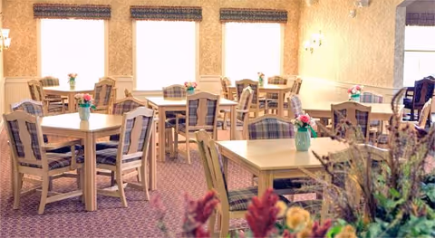 A bright dining room with multiple wooden tables and chairs featuring plaid cushions. Each table has a small vase with flowers. Large windows with valances allow natural light to fill the room. The carpet is patterned, and there are floral arrangements in the foreground.
