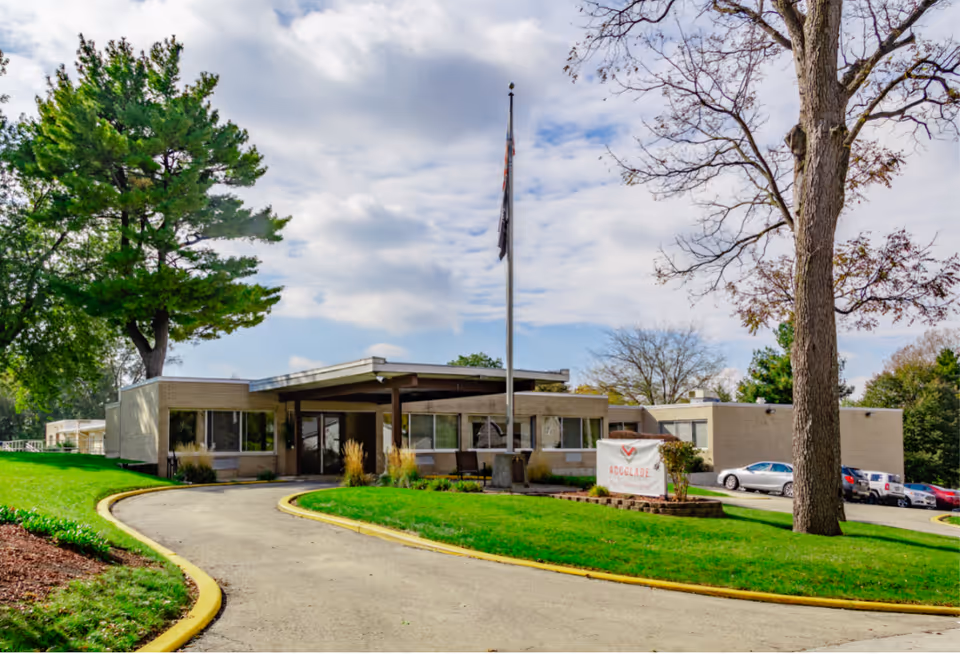 Exterior view of Accolade Healthcare Of Paxton Senior Living building with a curved driveway, green lawn, trees, and a flagpole in front of the entrance under a partly cloudy sky.
