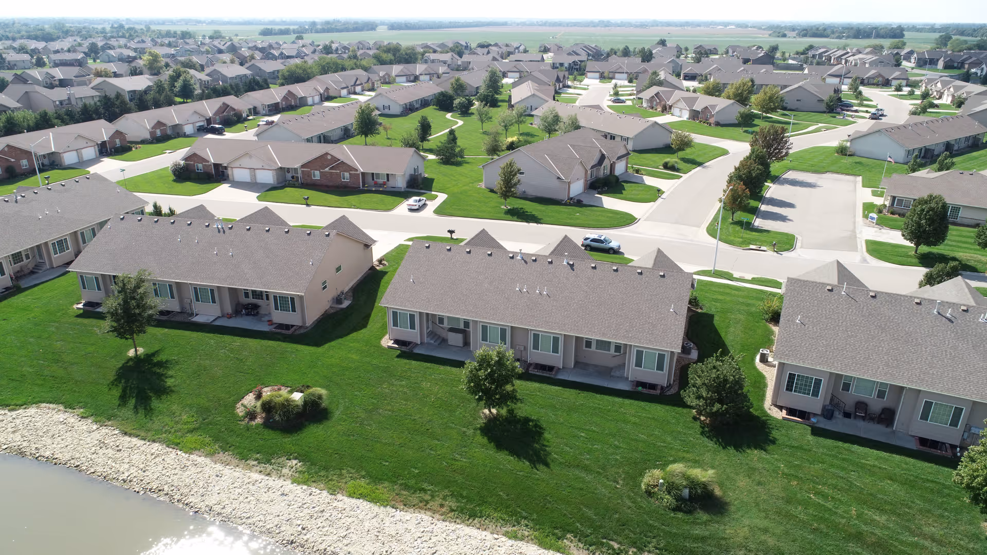 Aerial view of single-story residential villas with green lawns and a small pond in a suburban neighborhood.