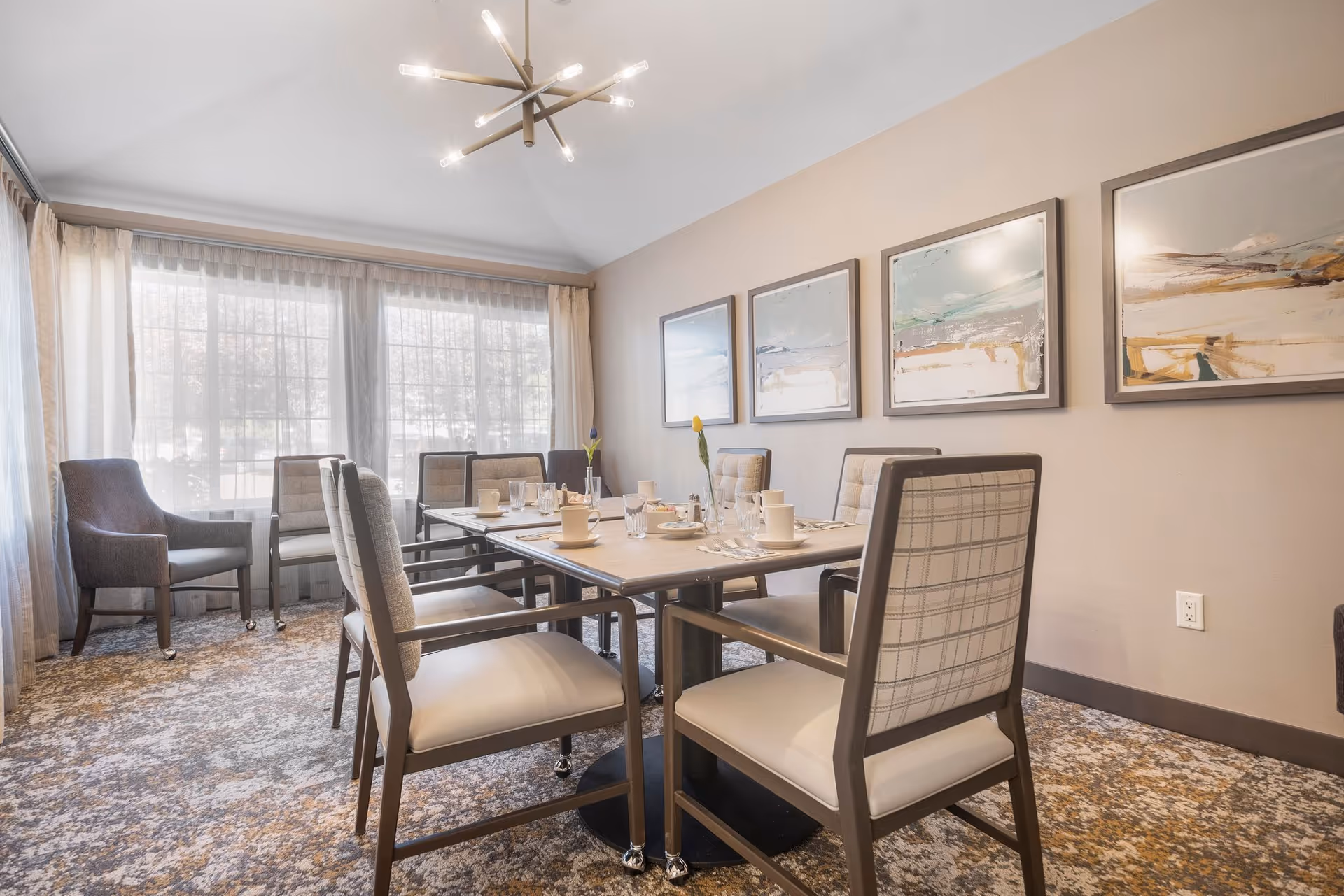 Bright dining room with a table set for several people, upholstered chairs, large windows with sheer curtains, and framed artwork on the wall.