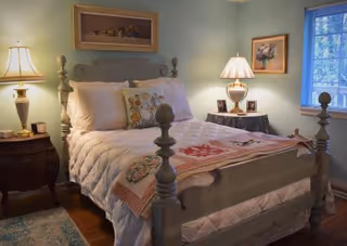 A cozy bedroom with a wooden bed frame painted in light gray, a white quilted bedspread, and decorative pillows. There are two bedside tables with lamps on each side of the bed. The walls are painted light blue, and there are framed pictures hanging on the walls. A window with blinds is visible on the right side.
