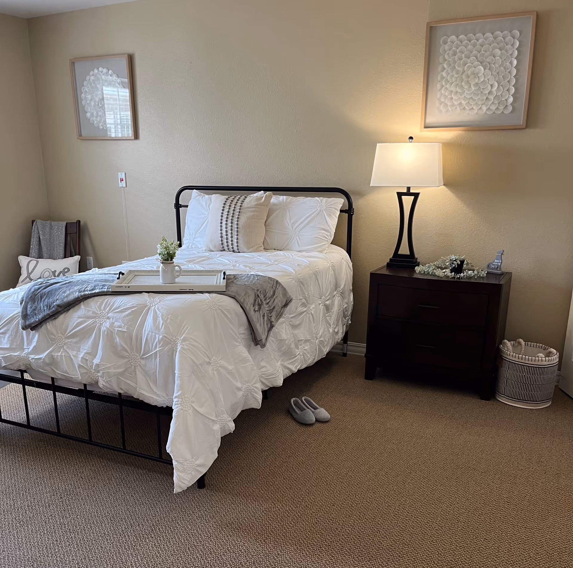 A neatly made bed with white bedding and decorative pillows in a bedroom. A tray with a small plant is placed on the bed. To the right of the bed is a dark wooden nightstand with a lamp, decorative items, and a basket on the floor beside it. On the left side, there is a chair with a gray blanket and a pillow with the word 'love'. Two framed artworks hang on the beige walls above the bed and nightstand. A pair of gray slippers is on the carpeted floor near the bed.