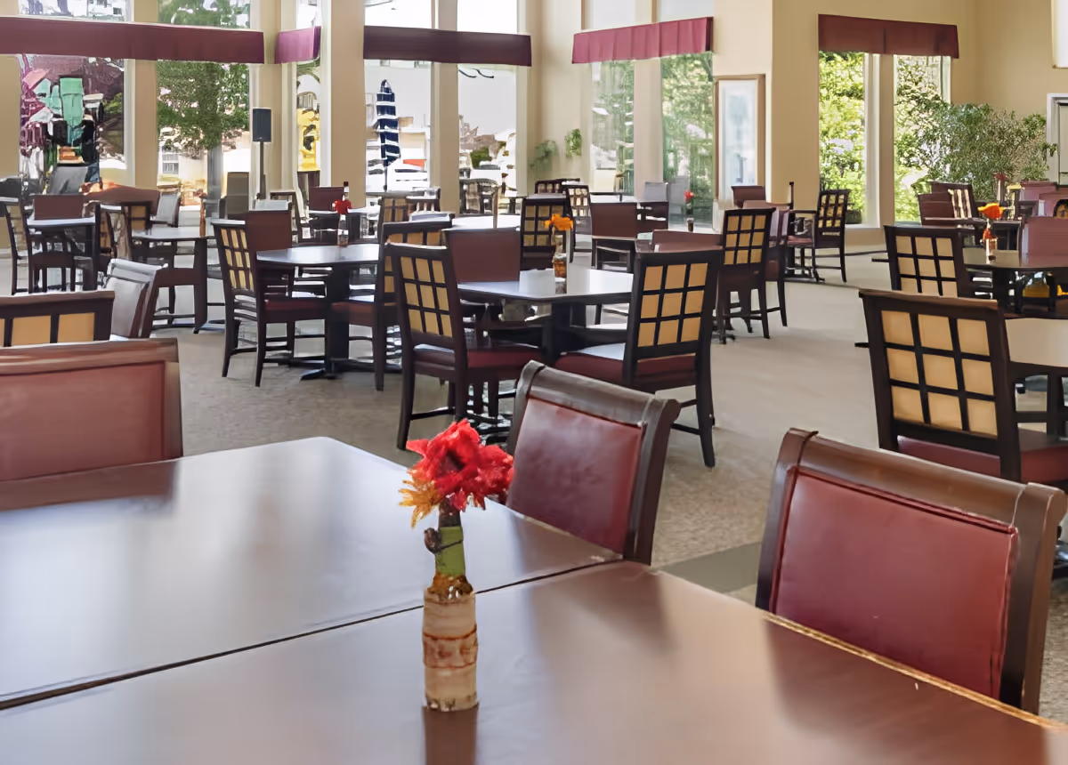 Spacious dining room with multiple tables and chairs, large windows, and a small flower vase on a foreground table.
