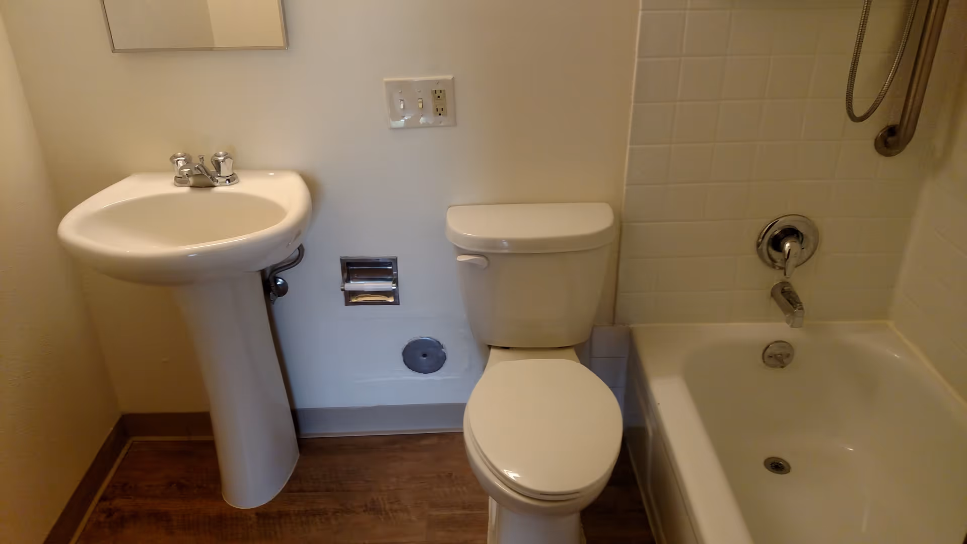 A small bathroom with a white pedestal sink, a toilet, and a bathtub with a showerhead and grab bar. The walls are light-colored, and the floor has a wood-like finish.