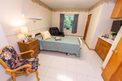 A simple, clean bedroom in a skilled nursing facility featuring a single bed with a light blue bedspread, a wooden nightstand with a lamp and picture frame, a floral upholstered armchair, a black recliner near a window with blue curtains, and wooden cabinets with a TV on top.