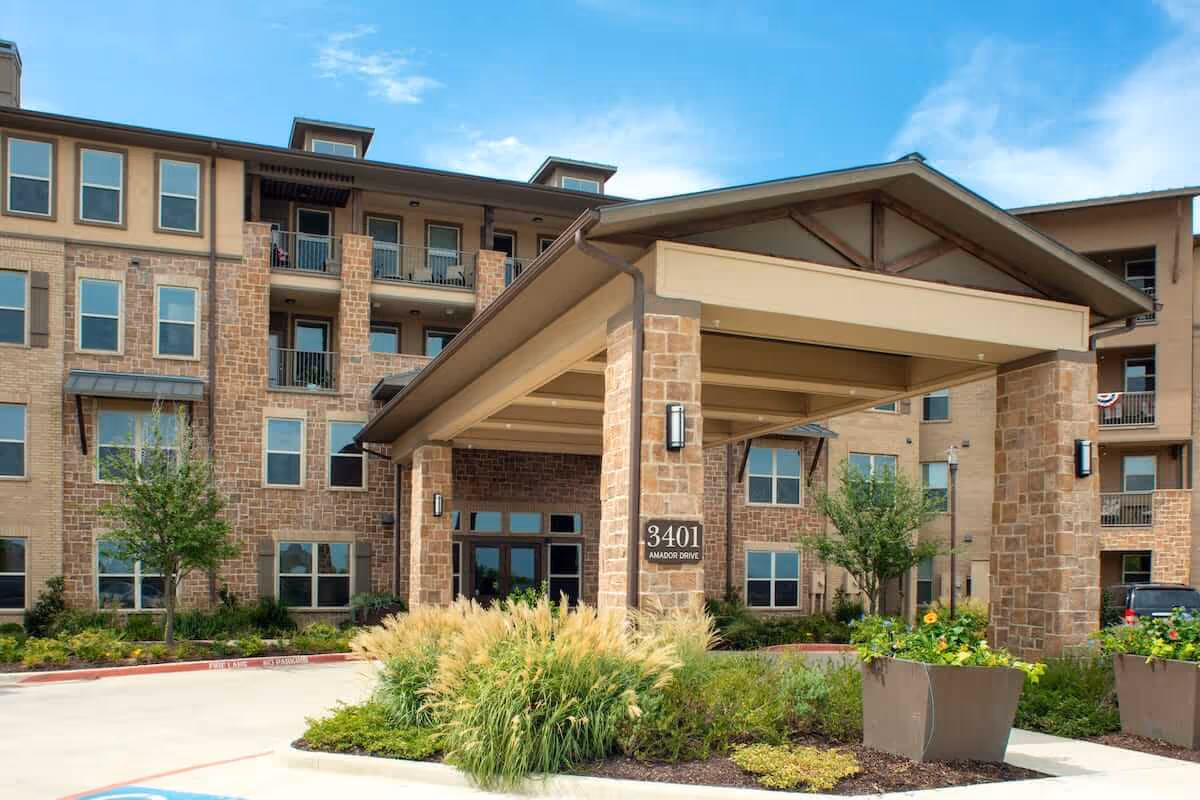 Exterior view of a multi-story senior living facility building with a covered entrance supported by stone pillars. The building has numerous windows and balconies, surrounded by landscaped greenery and plants under a blue sky.