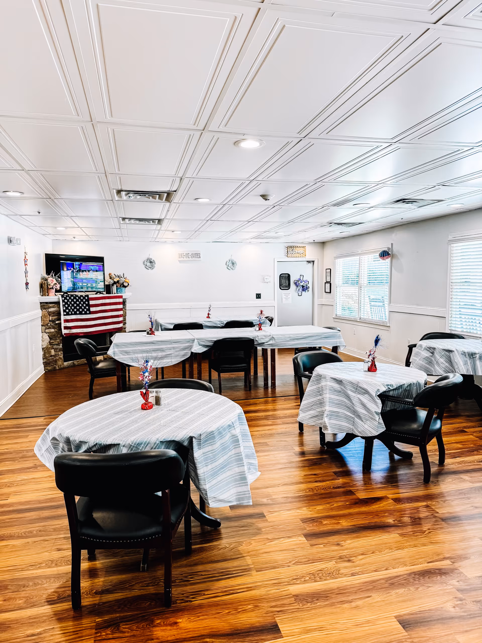 A dining room with several tables covered in light blue and white striped tablecloths, each table surrounded by black chairs. The room has wooden flooring, white walls, and a white paneled ceiling with recessed lighting. An American flag is draped over a stone fireplace in the corner, and a TV is mounted above it. Windows with blinds allow natural light into the room.