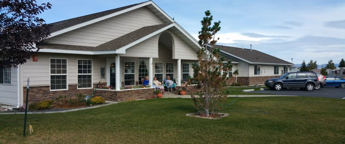 Exterior view of a single-story assisted living facility building with a covered porch where three elderly people are seated. The building has beige siding with stone accents and multiple windows. There is a well-maintained lawn with a small tree in the foreground and a parking lot with several cars to the right.