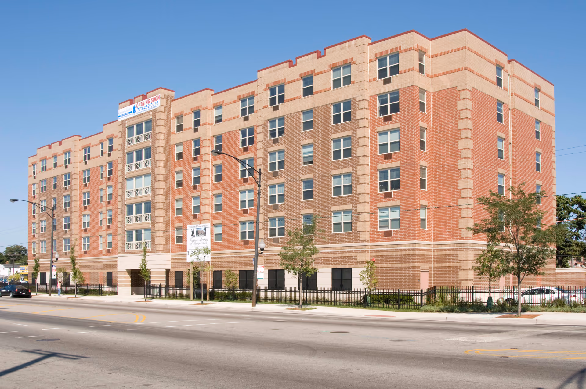 Exterior view of a multi-story brick senior living facility building with multiple windows, a fenced area, and a clear blue sky.