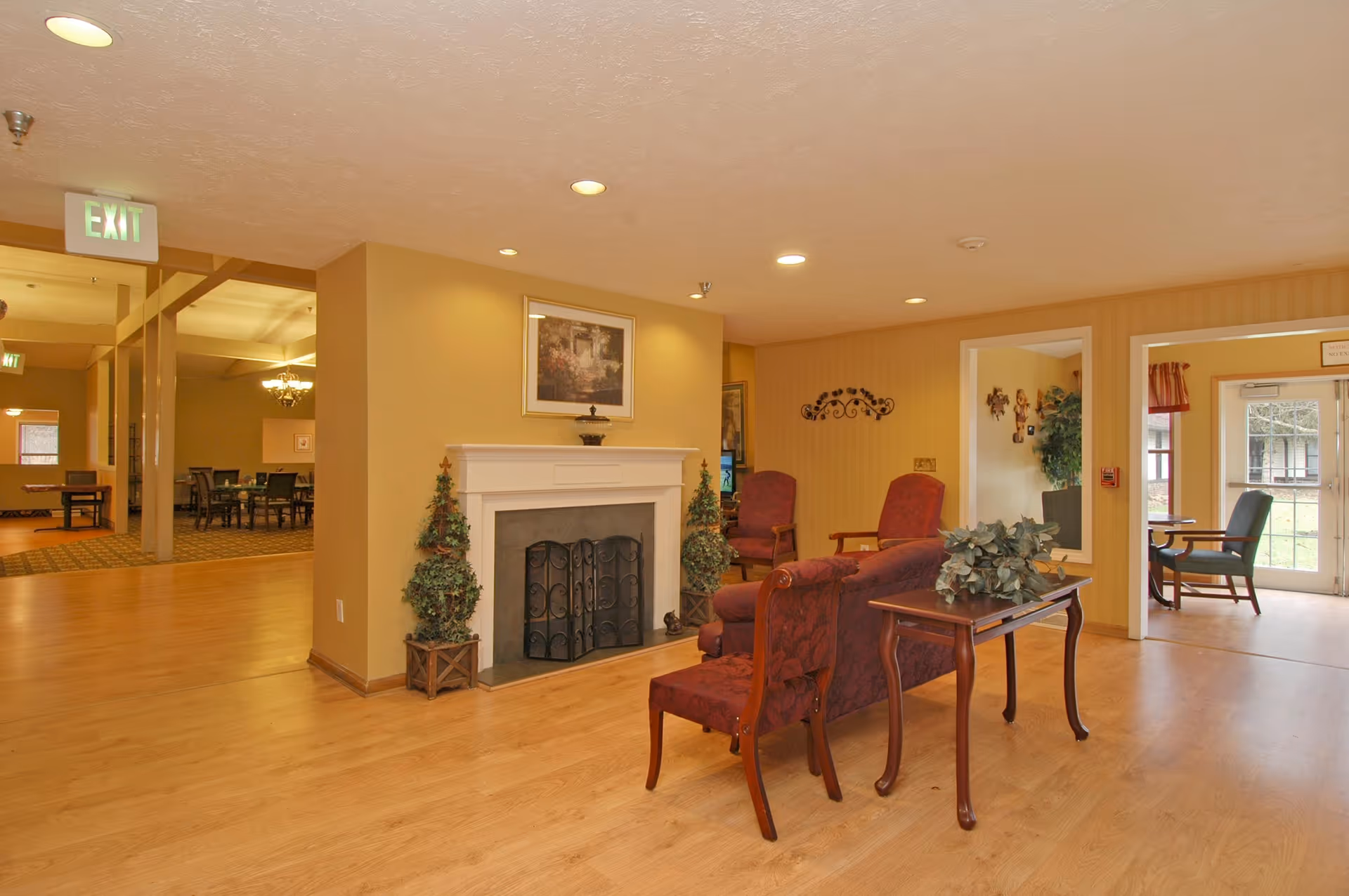 A cozy living room area in a senior living facility featuring a fireplace with decorative plants on either side, a framed picture above the mantel, and comfortable upholstered chairs and a small table with a plant. The room has warm yellow walls and wood flooring, with an adjacent dining area visible in the background and a glass door leading outside.