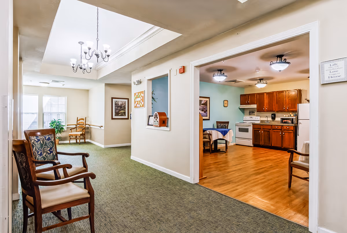 Interior view of a senior living facility showing a hallway with carpeted floor and wooden chairs with patterned cushions. To the right, there is an open kitchen area with wooden cabinets, a white stove, refrigerator, and a small dining table with a blue and white tablecloth. The hallway has beige walls, a skylight with a chandelier, and framed artwork on the walls.