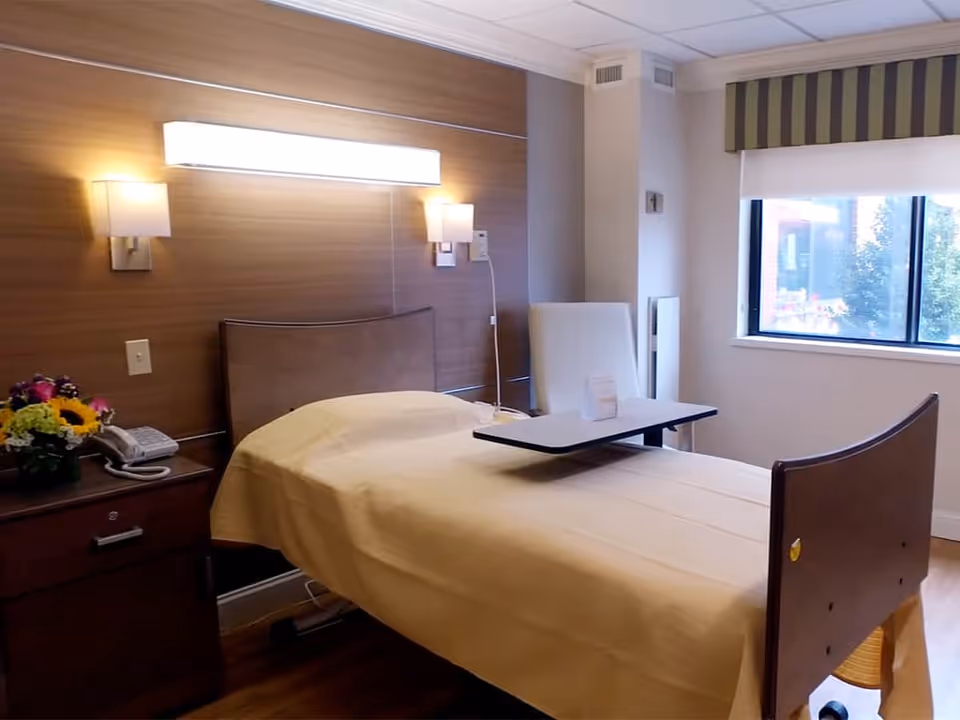 A neatly made single bed with a beige blanket in a senior living facility room. The bed has a movable overbed table and is positioned next to a wooden nightstand with a telephone and a vase of flowers. The room features a large window with a striped valance, a white armchair, and warm wall lighting above the bed.