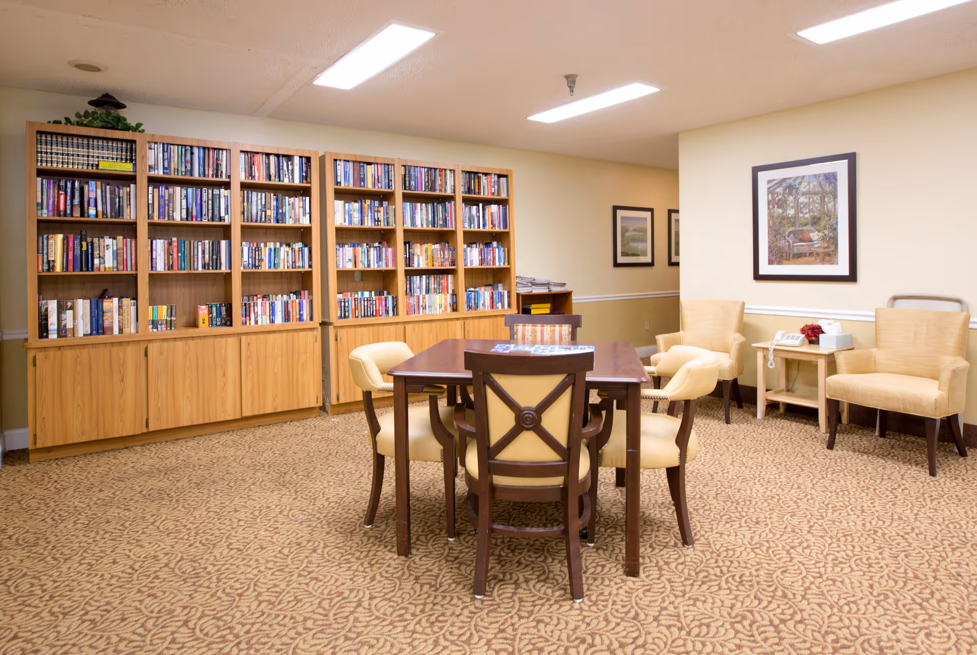 A communal reading room with bookshelves along the back wall, a central table surrounded by chairs, and armchairs against a beige wall.