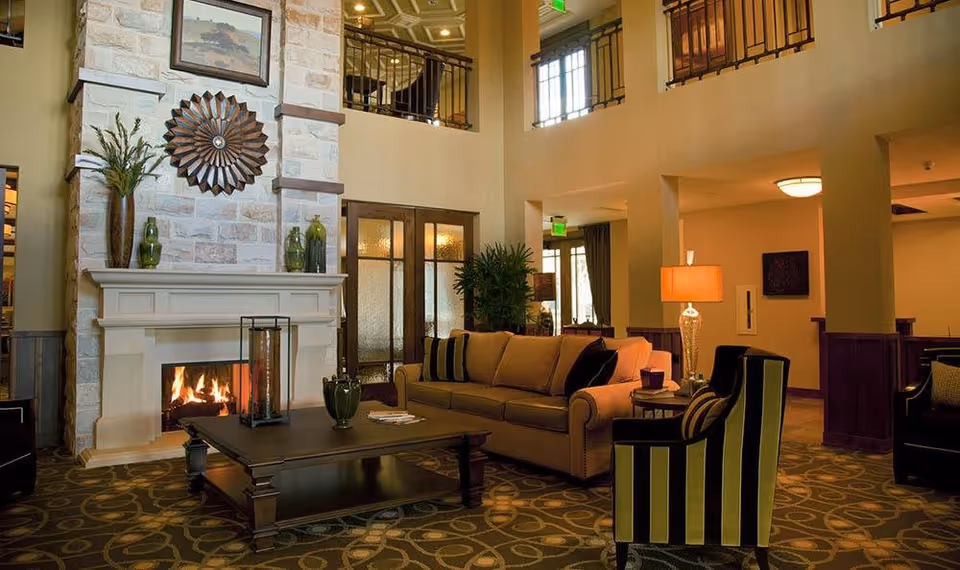 A cozy living room area with a lit fireplace set in a stone mantel. There is a beige sofa with black and beige striped pillows, a matching striped armchair, a dark wooden coffee table with decorative items, and a side table with a lamp. The room has warm lighting, a patterned carpet, and a second-floor balcony railing visible above.