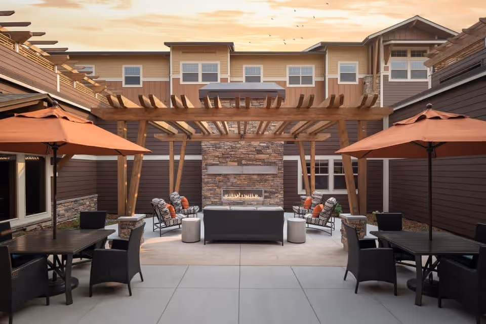 Outdoor patio area at The Springs at Bozeman featuring a stone fireplace with a wooden pergola overhead, surrounded by cushioned chairs and sofas. There are two tables with orange umbrellas and black chairs on either side of the patio. The building exterior is visible in the background under a partly cloudy sky at sunset.