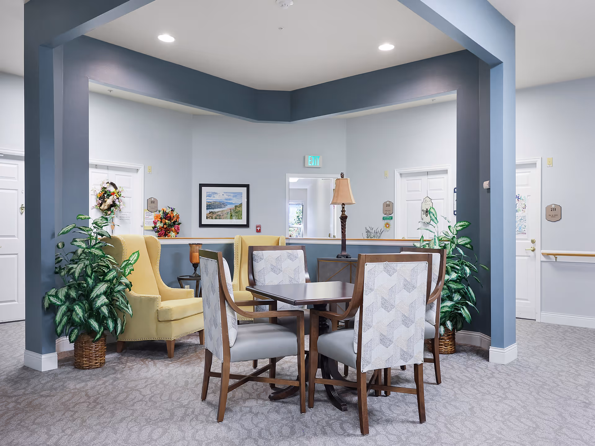 A cozy seating area in a senior living facility with a square wooden table surrounded by four cushioned chairs with patterned backs. Behind the table are two yellow upholstered armchairs and a wooden side table with a lamp. The walls are light gray with white doors and decorative wreaths. There are green potted plants on either side of the seating area, and a framed landscape painting hangs on the wall.