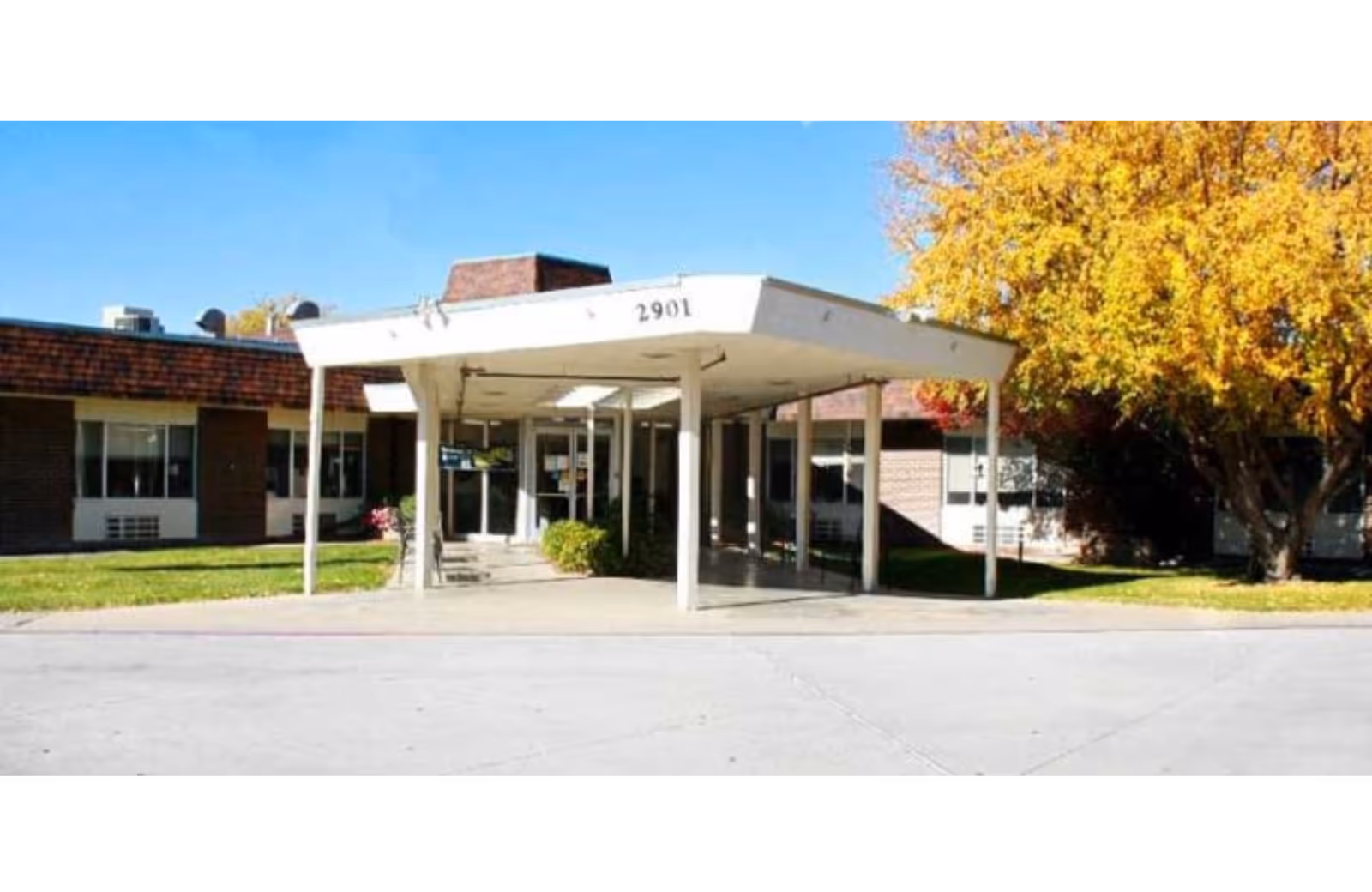 Front entrance of a single-story brick care facility with a covered drive-up canopy marked "2901" and autumn trees nearby.