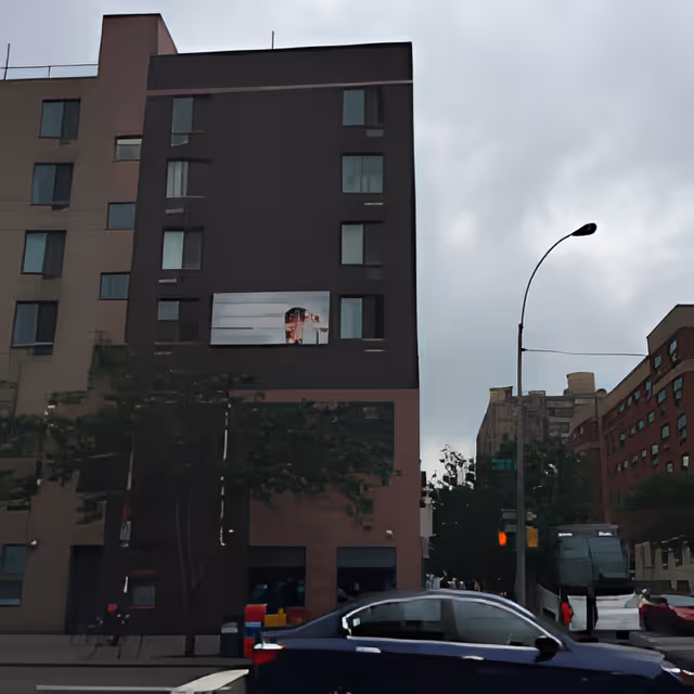Exterior view of a multi-story building on a city street with several windows, a tree in front, a streetlight, and a dark blue car driving by. The sky is cloudy.