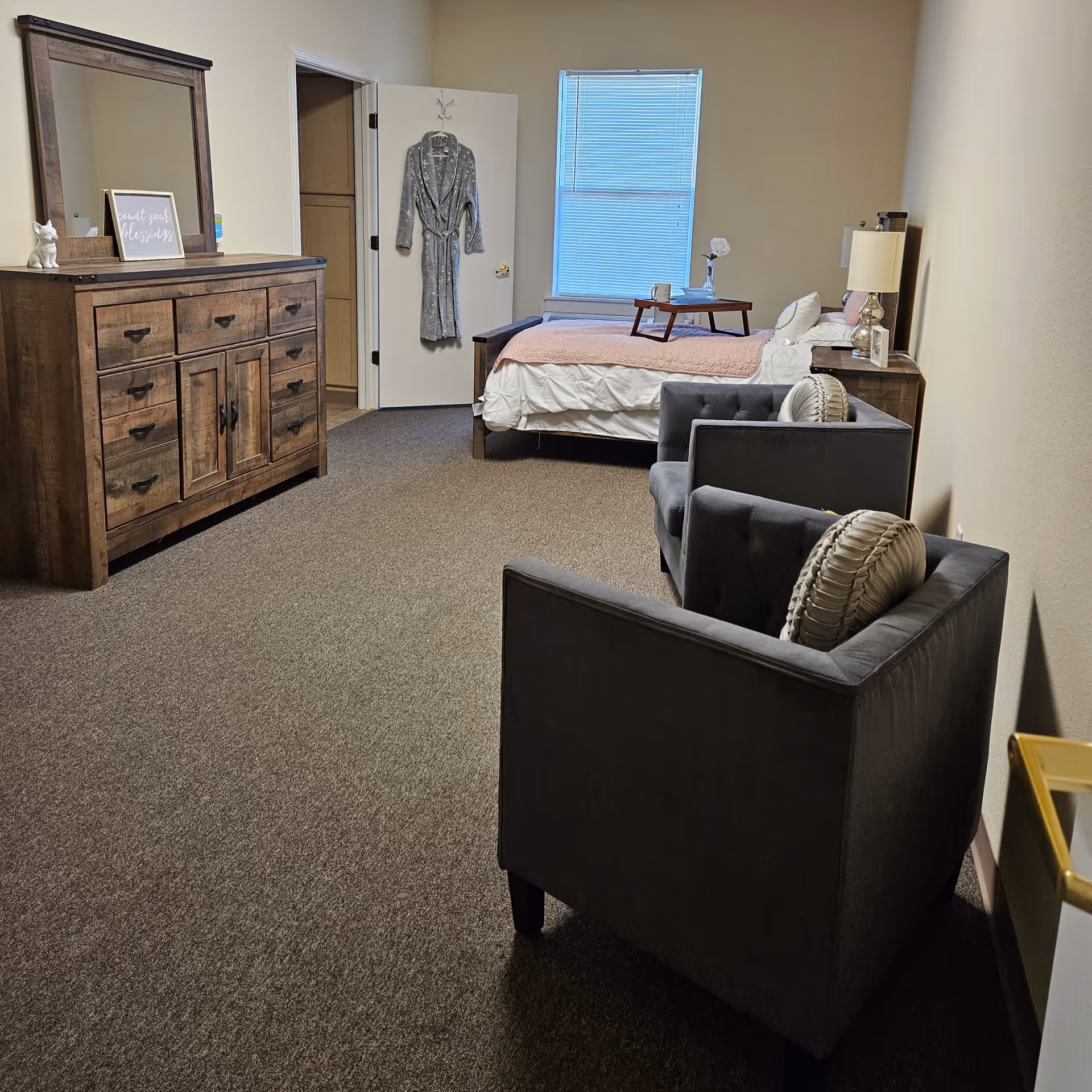 A senior living bedroom with a bed covered in white and pink bedding, a wooden nightstand with a lamp and decorative items, two gray armchairs with round pillows, a wooden dresser with a mirror, and a door with a gray robe hanging on a hook. The room has beige walls and carpeted floor.