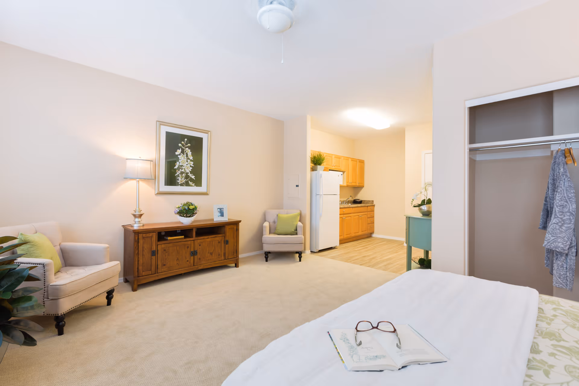 A bright and cozy senior living studio apartment featuring a bed with an open book and glasses on it, an open closet with hanging clothes, two beige armchairs with green cushions, a wooden cabinet with a lamp, plant, and framed photo, and a small kitchen area with wooden cabinets, a white refrigerator, and a countertop.