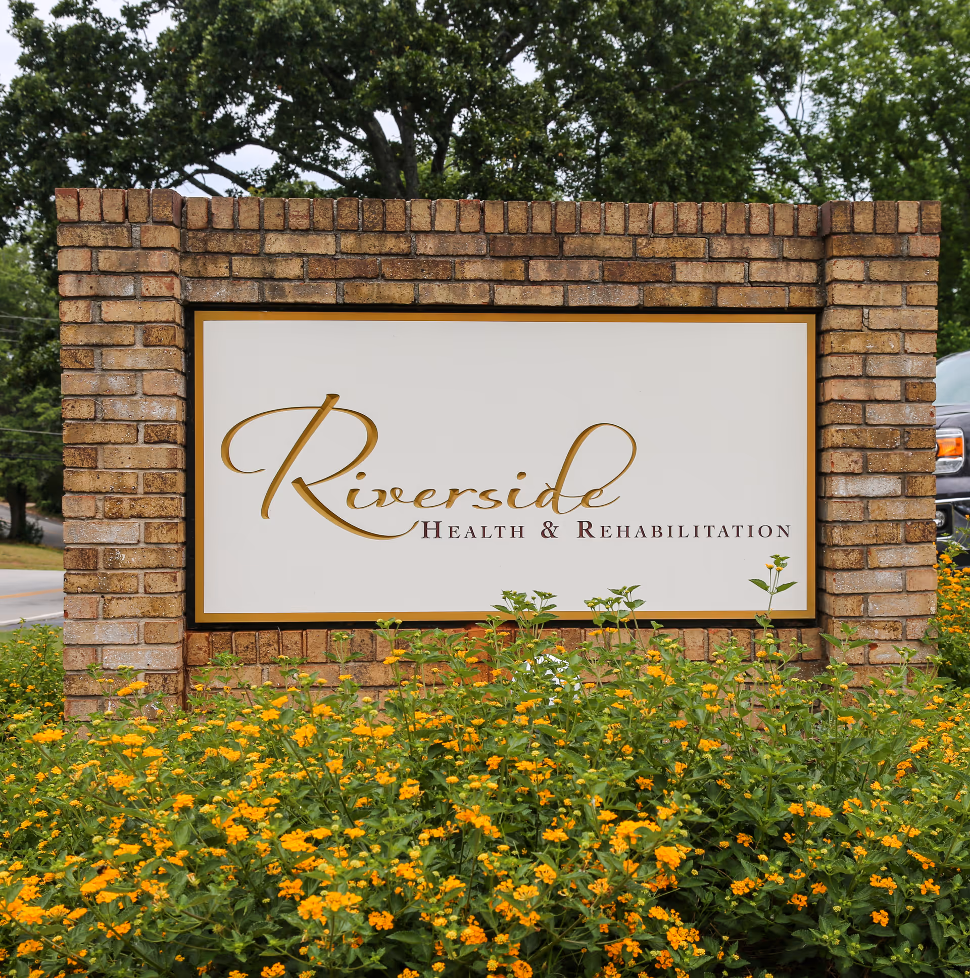 A brick sign for Riverside Health & Rehabilitation surrounded by green plants with small yellow flowers, with trees and a road visible in the background.