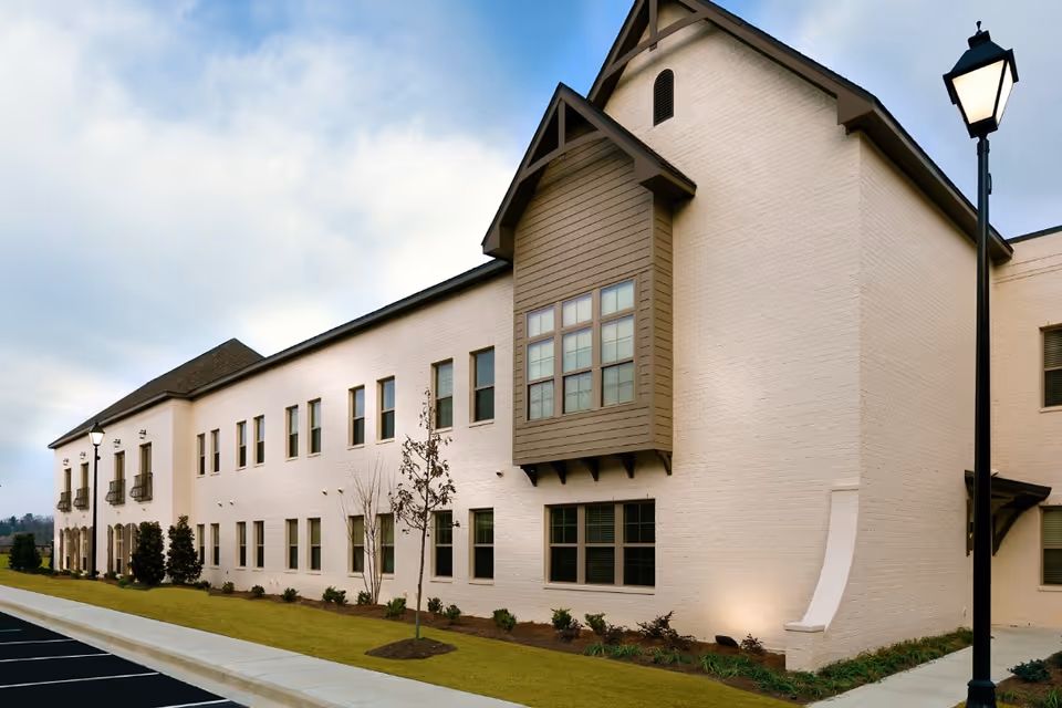 Exterior view of a two-story senior living facility building with beige brick walls, multiple windows, small landscaped bushes and trees, a sidewalk, and a street lamp on the right side under a partly cloudy sky.
