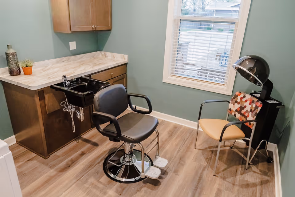 A small salon room with a black salon chair in front of a black sink attached to a wooden cabinet with a marble countertop. There is a window with white blinds on the wall, and next to it is a chair with a colorful hexagonal patterned backrest and a beige seat. A black hair dryer hood is positioned above the chair. The room has light wood flooring and green walls.