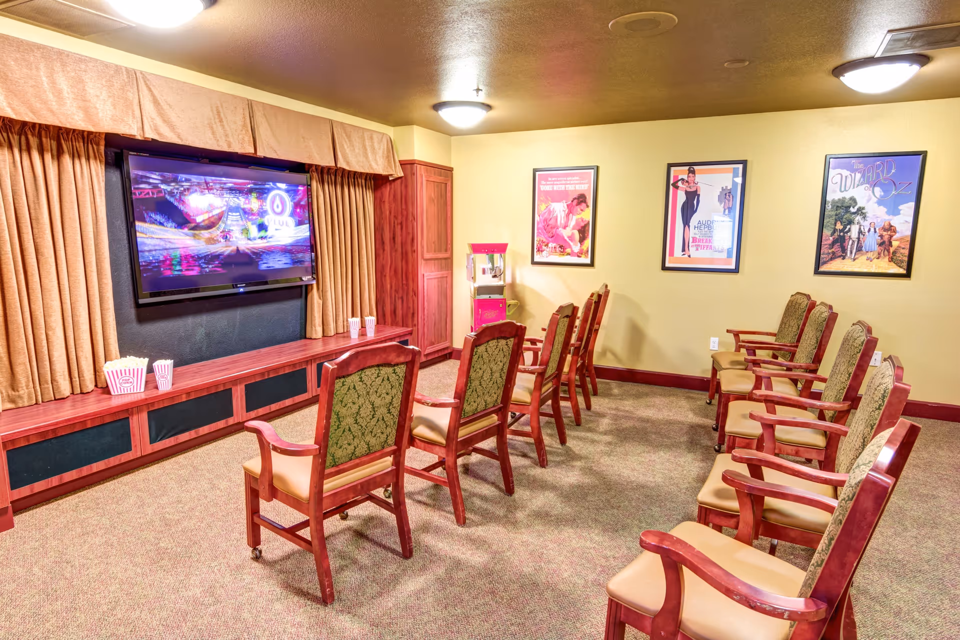 A small theater room with two rows of wooden chairs with green patterned backs and beige seats facing a wall-mounted flat screen TV. There are three framed movie posters on the yellow wall to the right and a popcorn machine in the corner. Popcorn containers are placed on the wooden ledge beneath the TV.