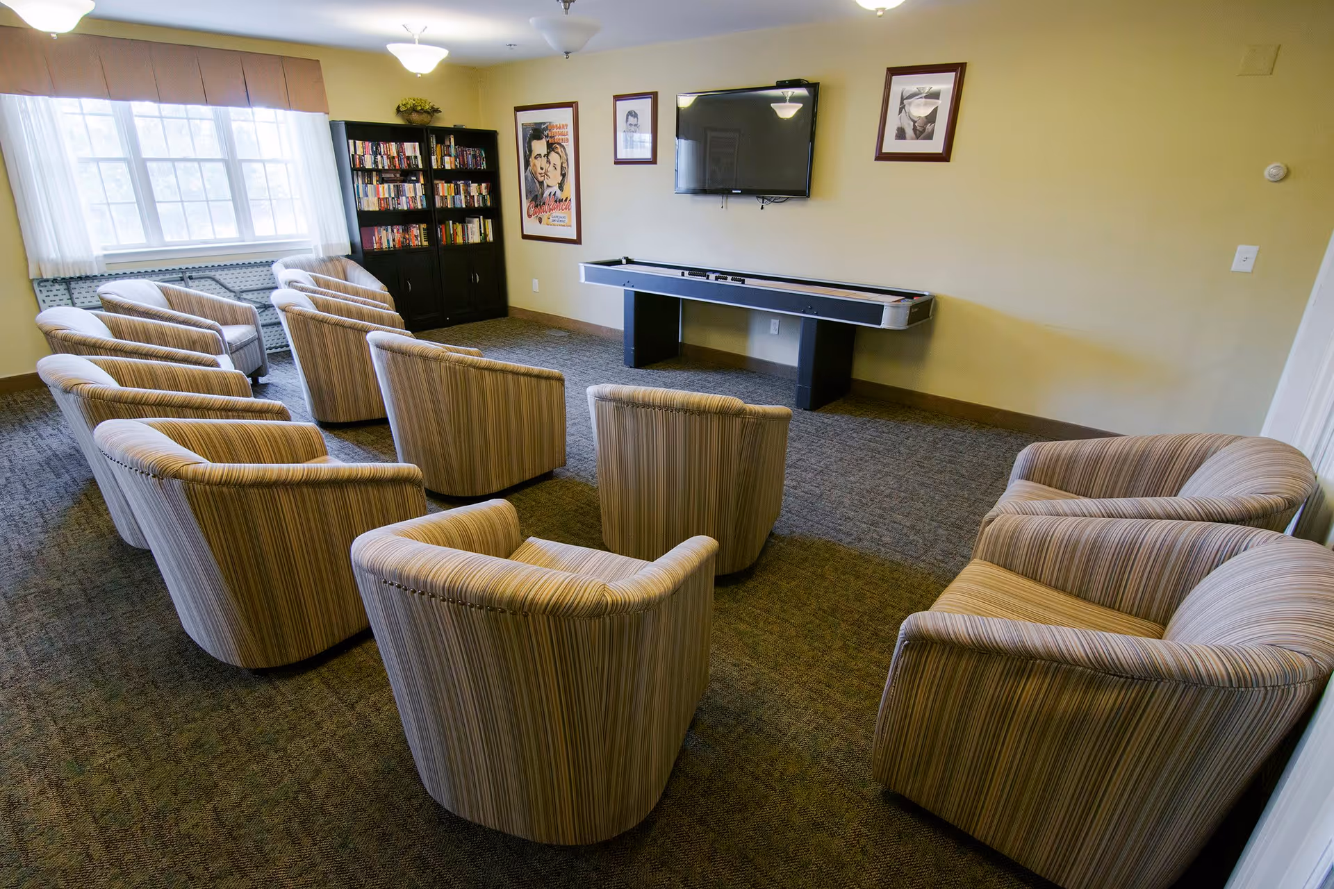 A senior living facility common room with multiple striped armchairs arranged in rows facing a wall-mounted flat screen TV and a shuffleboard table. There is a large window with sheer white curtains and a valance, a black bookshelf filled with books, and framed pictures on the walls.
