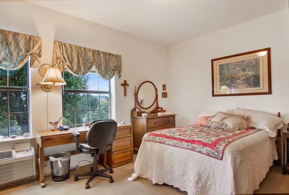 A cozy bedroom with a bed covered in a white quilt and a colorful patchwork blanket. There are several pillows on the bed. Next to the bed is a wooden nightstand with a lamp. A wooden dresser with a round mirror and some decorative items is against the wall. A wooden desk with a black office chair is positioned in front of two large windows with patterned valances. A wall-mounted lamp is above the desk, and an air conditioning unit is below the windows. The room has light-colored walls and carpeted floor.