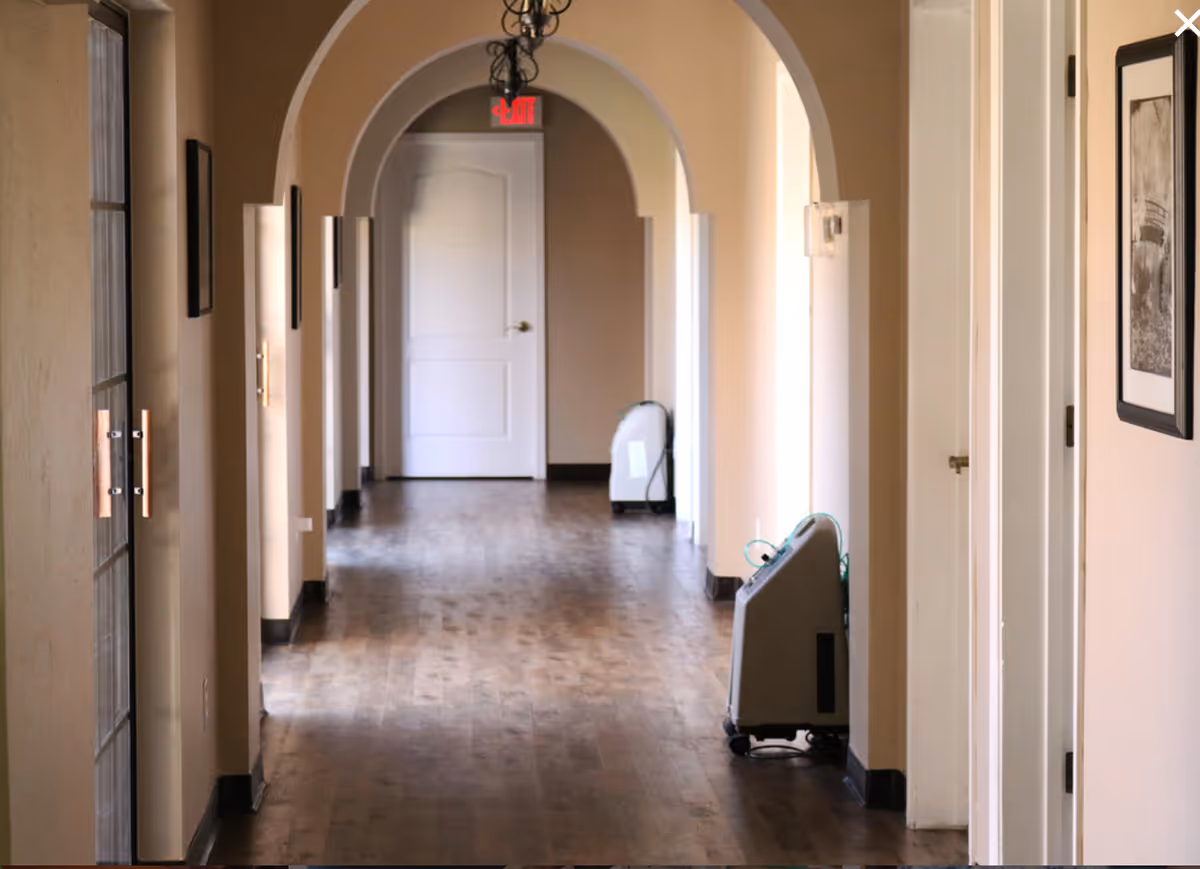 A long indoor hallway with wooden flooring and beige walls featuring multiple archways. There are framed pictures on the walls and two portable oxygen concentrators placed along the right side of the hallway. A white door with an illuminated red exit sign above it is visible at the end of the corridor.