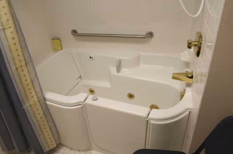 A walk-in bathtub with a door for easy access, equipped with gold-colored faucet and handles, a grab bar on the tiled wall, and a soap holder. The bathtub is designed for senior or disabled users to safely enter and exit.