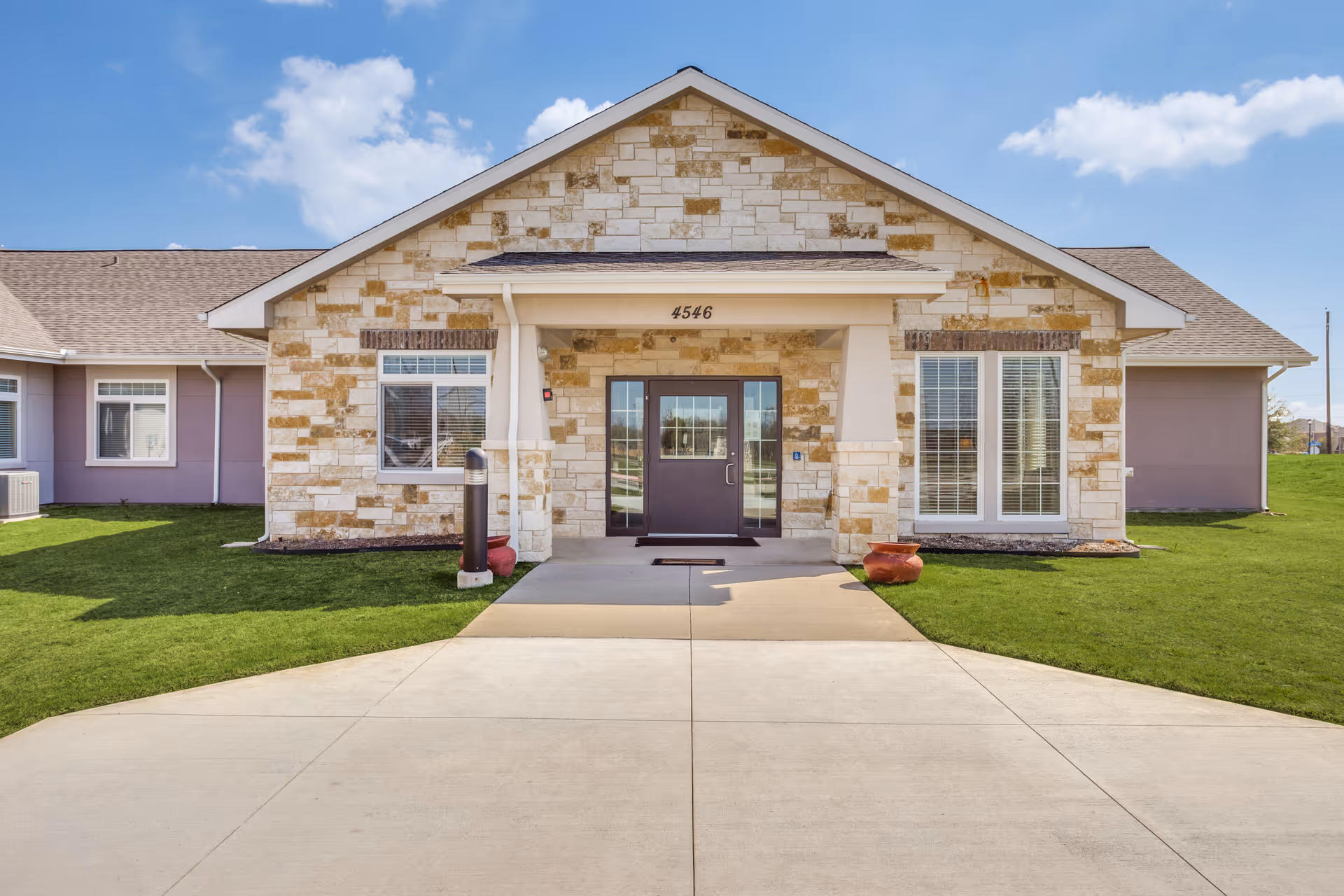 Front exterior view of a single-story building with a stone facade and a central entrance door under a small covered porch. The building is surrounded by green grass and a clear blue sky with some clouds.