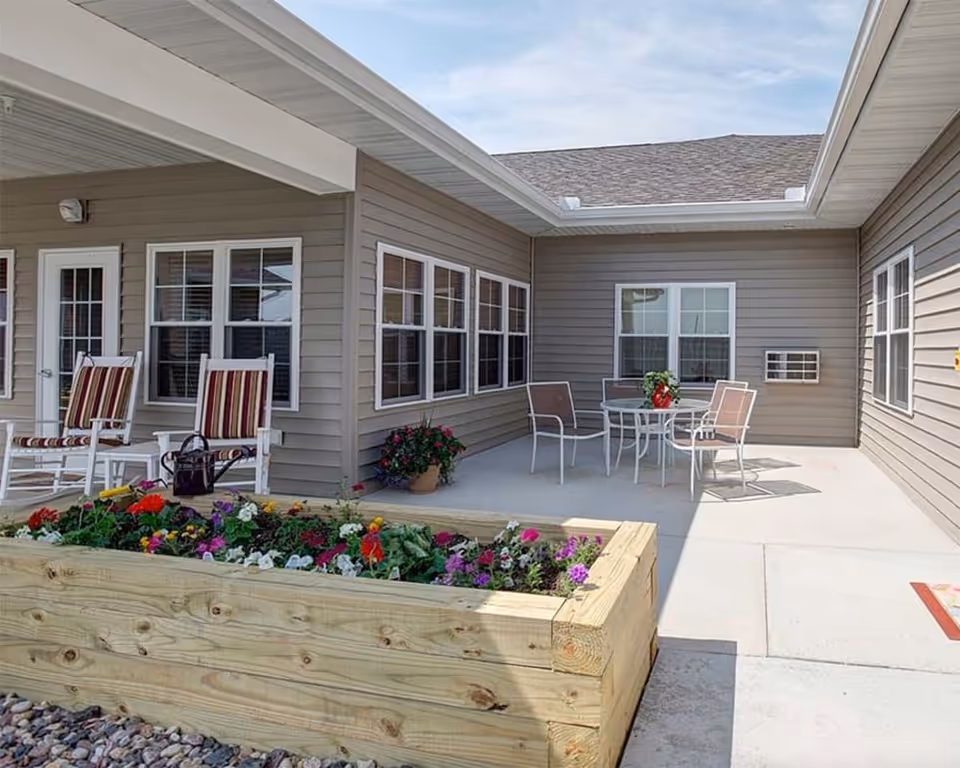 Outdoor patio area at Milestone Senior Living Hillsboro featuring a raised wooden flower bed with colorful flowers, two striped cushioned rocking chairs, a small white side table, and a round glass table with four chairs. The patio is surrounded by beige siding walls with multiple windows and a door.