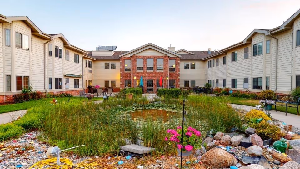 Outdoor courtyard area of a senior living facility with a pond surrounded by tall grasses and rocks in the center. The courtyard is enclosed by a two-story building with beige siding and brick accents. There are benches, patio tables with umbrellas, and landscaped greenery around the pond.