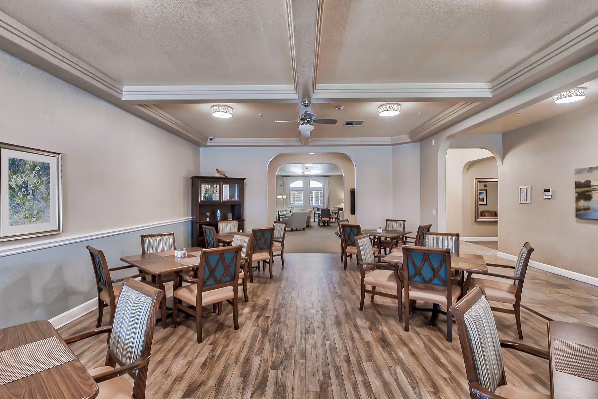 A spacious dining room with several wooden tables and chairs arranged neatly on a wood-patterned floor. The room has beige walls with framed artwork, a ceiling fan, and recessed lighting. In the background, there is an adjoining room with more seating and large windows letting in natural light.