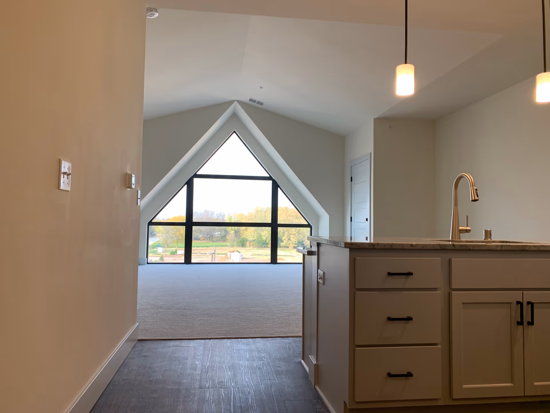 Interior view of a modern living space with a kitchen island featuring a sink and drawers on the right. The room has a large triangular window at the far end, letting in natural light and offering a view of trees and greenery outside. The floor transitions from dark wood near the kitchen to carpet in the living area. Two pendant lights hang from the ceiling above the kitchen island.