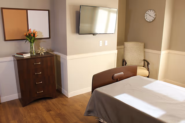 A tidy senior care bedroom with a made bed, wall-mounted TV, wooden dresser topped with flowers and books, and an armchair under a wall clock.