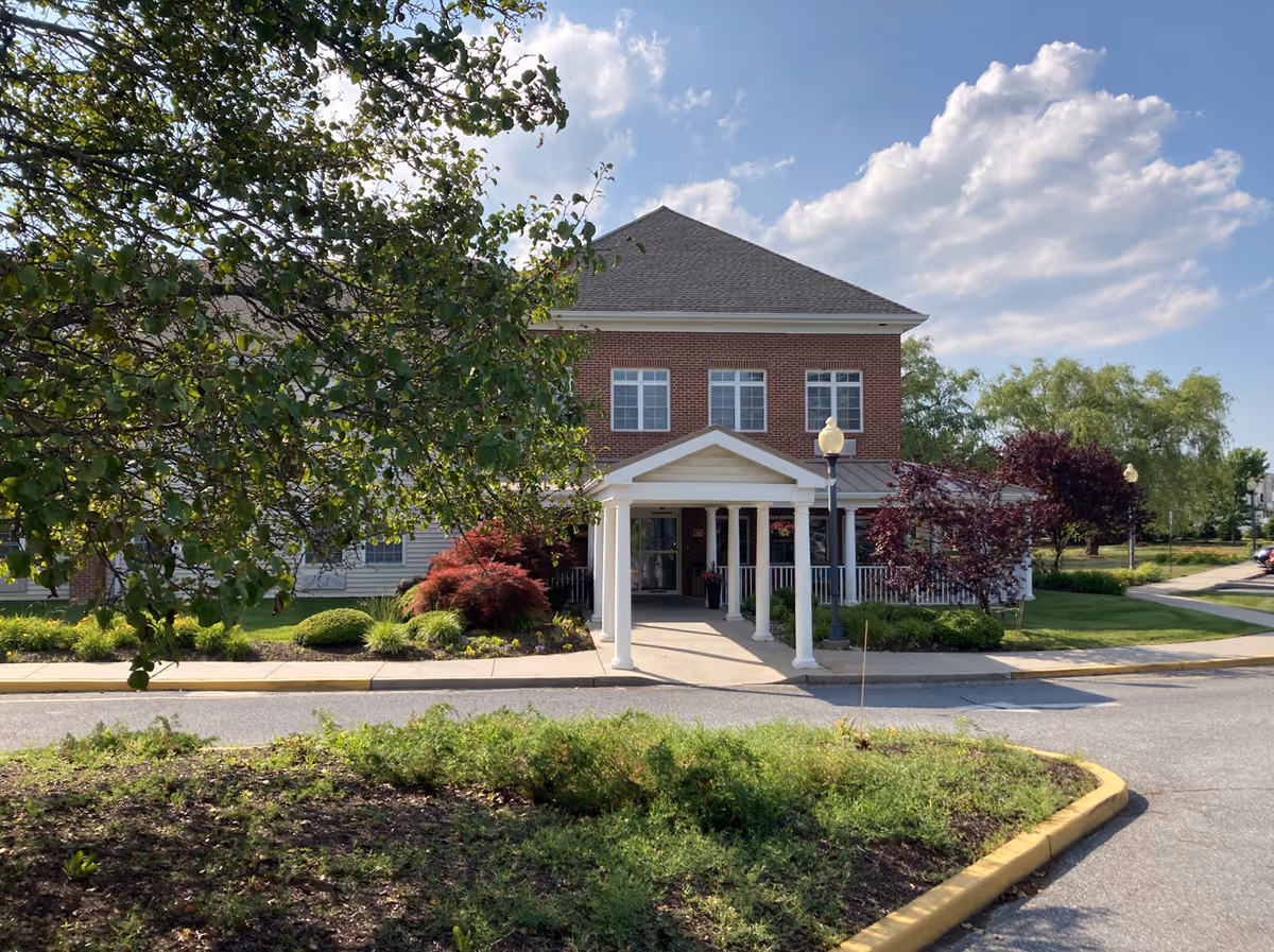 Front exterior of a two-story brick senior living building with a columned entrance, porch and surrounding trees and landscaping.