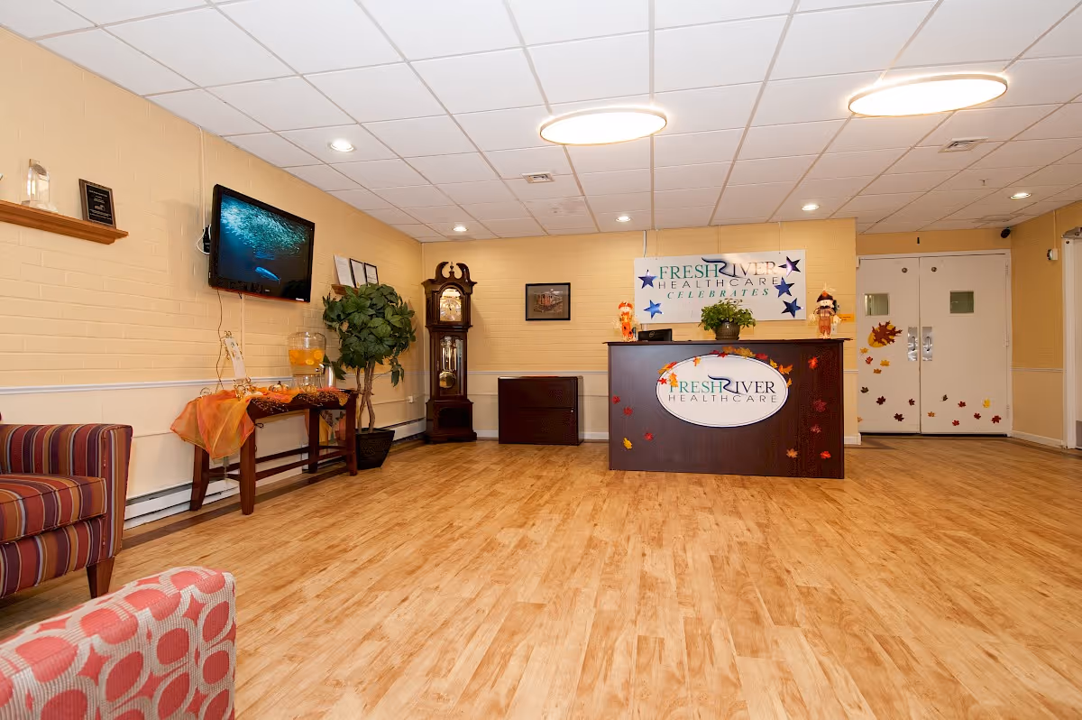 Reception area of Fresh River Healthcare, LLC with a wooden floor, a dark wood reception desk decorated with fall leaves, a wall-mounted TV, a grandfather clock, a potted plant, and seating with patterned upholstery.