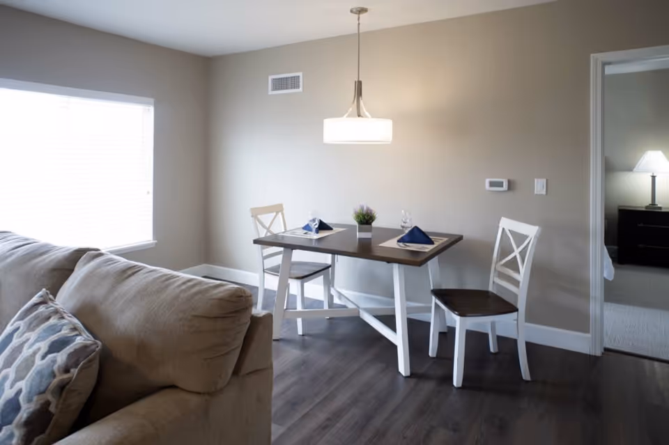 A small dining area with a wooden table set for two, featuring two white chairs with dark wooden seats, a small potted plant centerpiece, and two folded navy blue napkins on placemats. To the left, part of a beige sofa with a patterned pillow is visible. The room has light-colored walls, dark wood flooring, a large window with blinds, and a hanging light fixture above the table. A doorway on the right leads to a bedroom with a bedside table and lamp.