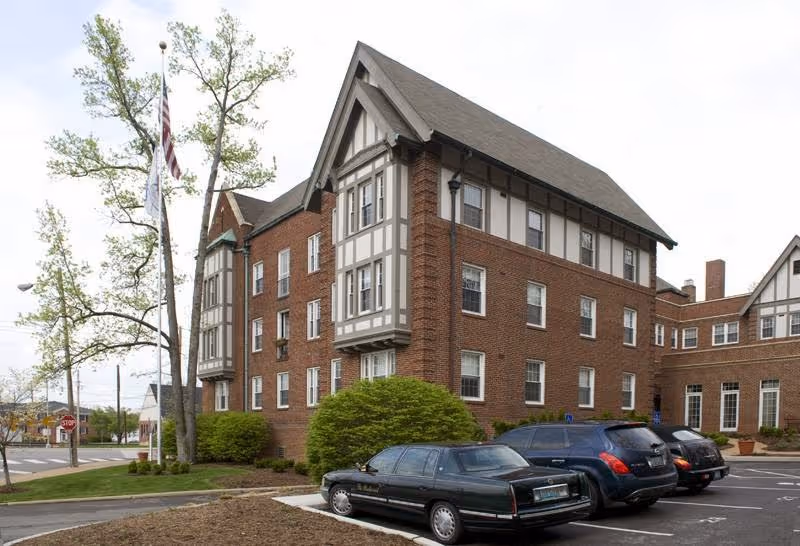 Exterior view of a multi-story brick building with Tudor-style architectural elements, surrounded by a parking lot with several parked cars and a flagpole with an American flag and another flag. Trees and shrubs are visible around the building.