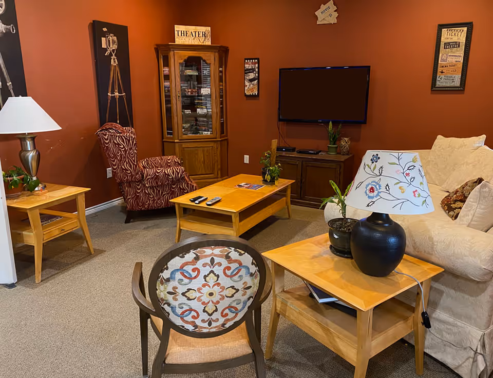 A cozy living room with a beige sofa, a wooden coffee table, a patterned armchair, and a wooden side table with a decorative lamp and a small plant. The walls are painted a warm reddish-brown color and decorated with framed theater-themed artwork. A flat-screen TV is mounted on the wall above a wooden cabinet.