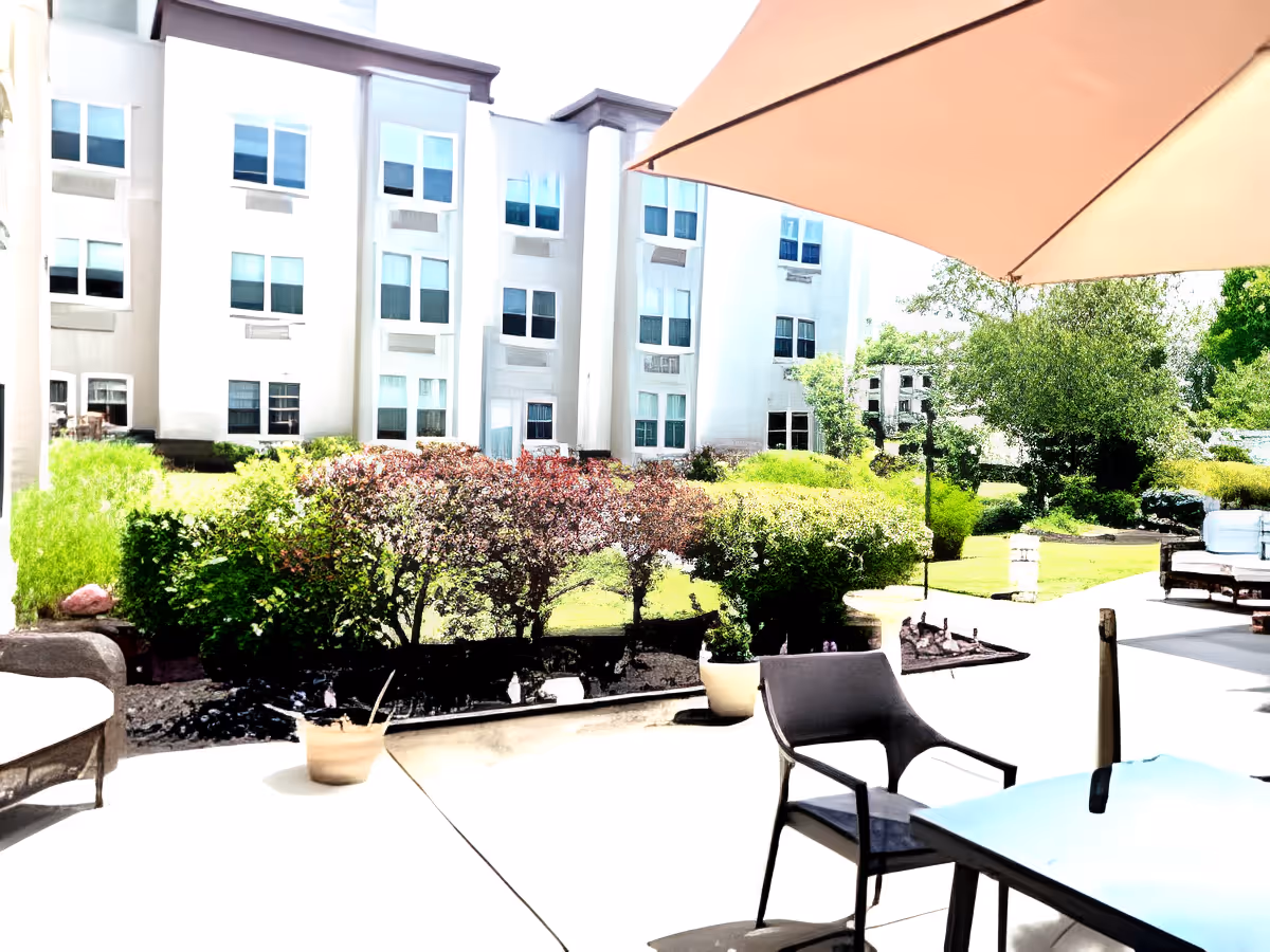 Outdoor patio area with a large umbrella, chairs, and tables. In the background, there is a multi-story white building with many windows and a well-maintained garden with bushes and trees.
