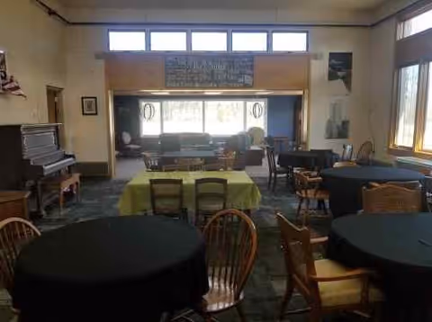 A spacious common area in a senior community with several round tables covered with black tablecloths and wooden chairs around them. There is a piano on the left side, large windows on the right letting in natural light, and a seating area with couches and chairs in the back near a large window. The room has a cozy and inviting atmosphere.