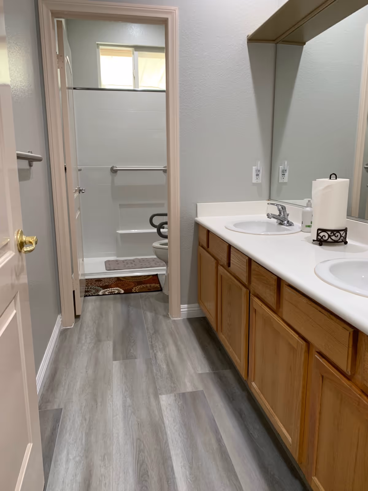 A clean bathroom with a double sink vanity featuring wooden cabinets and a large mirror. The floor is covered with gray wood-like tiles. In the background, there is a separate room with a toilet and a shower area with grab bars and a small window above.