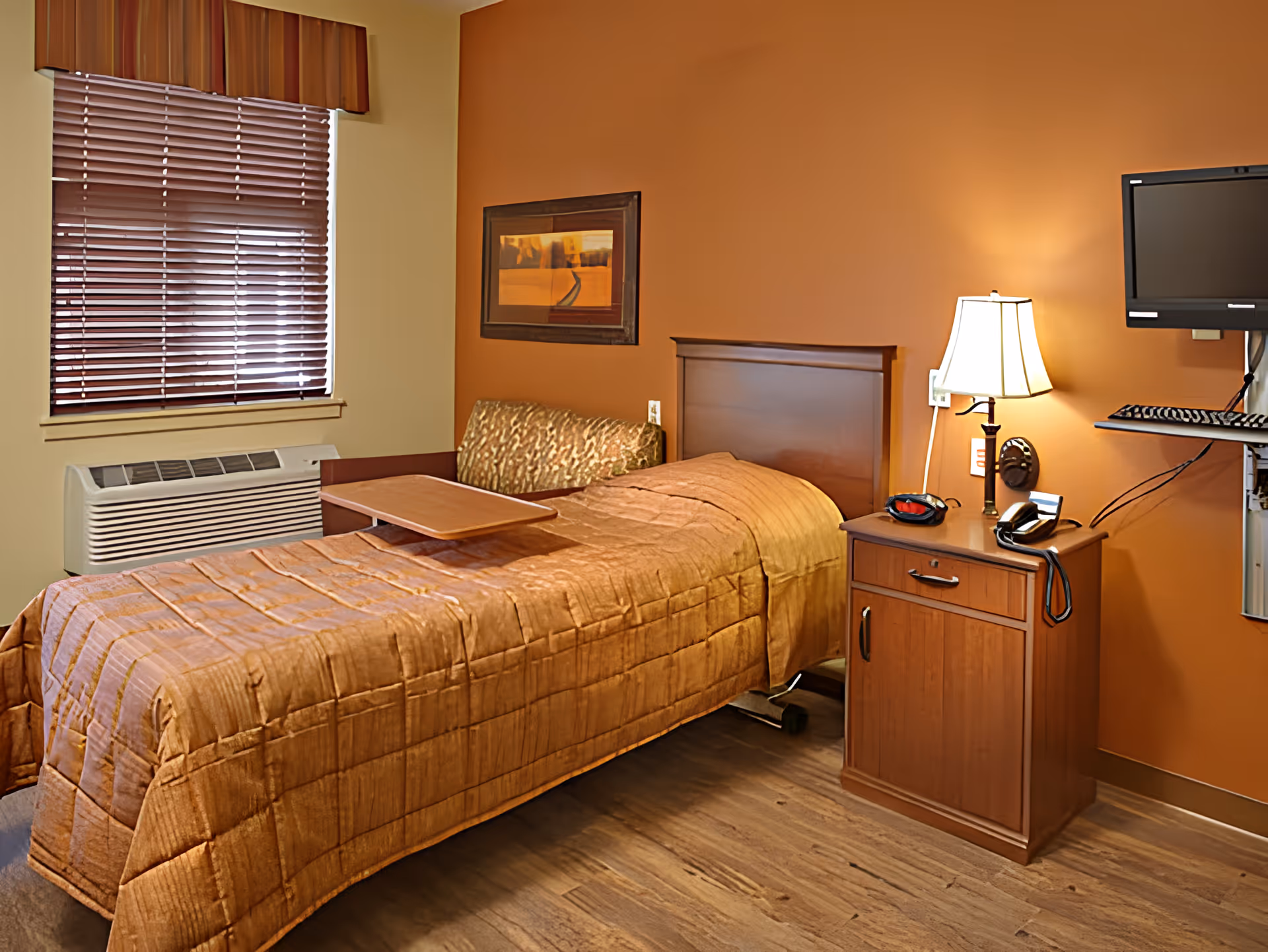 A single bed with a brown quilted bedspread in a room with orange walls. Next to the bed is a wooden nightstand with a lamp, telephone, and a mounted flat-screen TV above it. There is a window with closed blinds and a valance, and a small couch with a patterned cushion behind the bed.