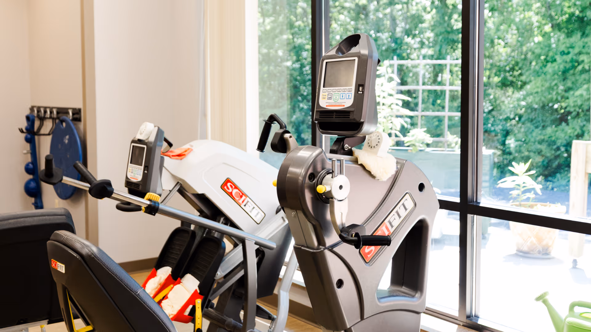 A bright exercise room with two SciFit exercise machines positioned near large windows that let in natural light and offer a view of greenery outside. The room has light-colored walls and a rack with exercise balls and other equipment in the background.