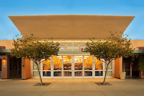 Front exterior view of Beatitudes Campus building entrance with large glass doors, two small trees in planters on either side, and a wide overhanging roof. The interior visible through the glass doors shows a well-lit dining area with tables and chairs.